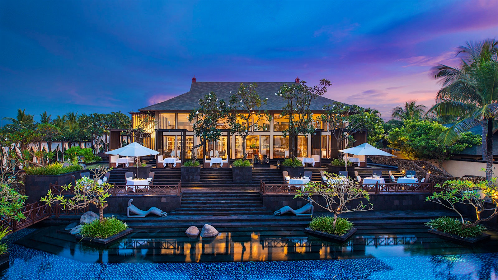 The bungalow-style exterior of Kayuputi restaurant at St. Regis Bali Resort at night, with a view of outdoor stairs and umbrellas